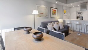 A modern living room with a grey sofa, a wooden dining table with two decorative bowls, and a kitchen area with white cabinets and a microwave. - 6-guest Haven Near Golden Mile (Madrid)