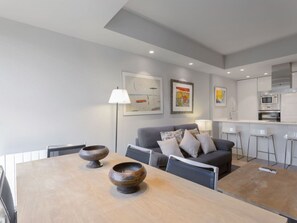 A modern living room with a grey sofa, a wooden dining table with two decorative bowls, and a kitchen area with white cabinets and a microwave. - 6-guest Haven Near Golden Mile (Madrid)