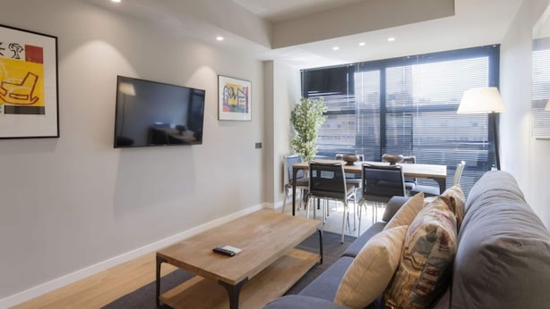 A modern living room with a sofa, a coffee table, a TV mounted on the wall, and a dining area with a table and chairs. - 6-guest Haven Near Golden Mile (Madrid)