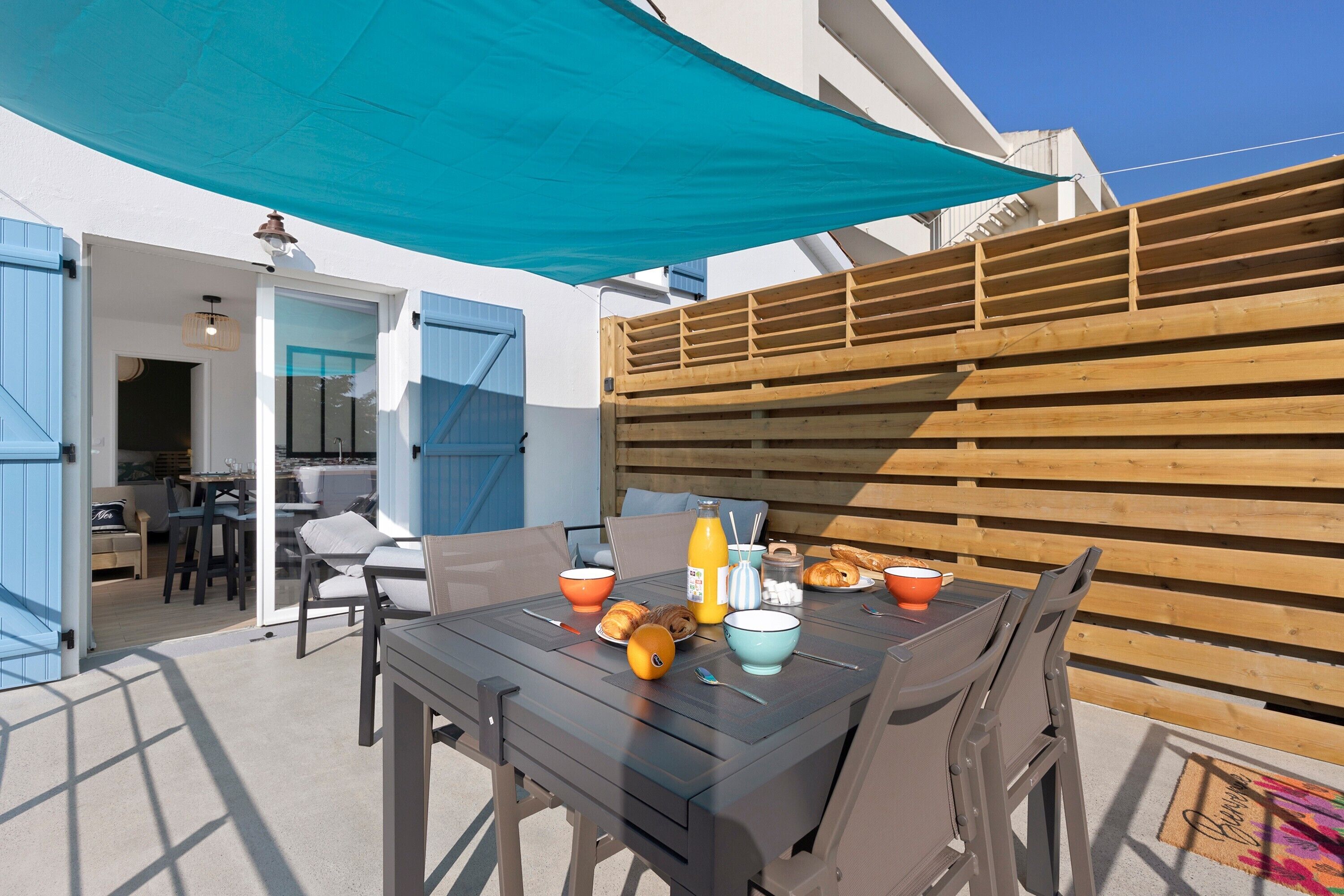 Un patio con una mesa dispuesta para el desayuno, incluyendo cruasanes y zumo de naranja, bajo un toldo azul.