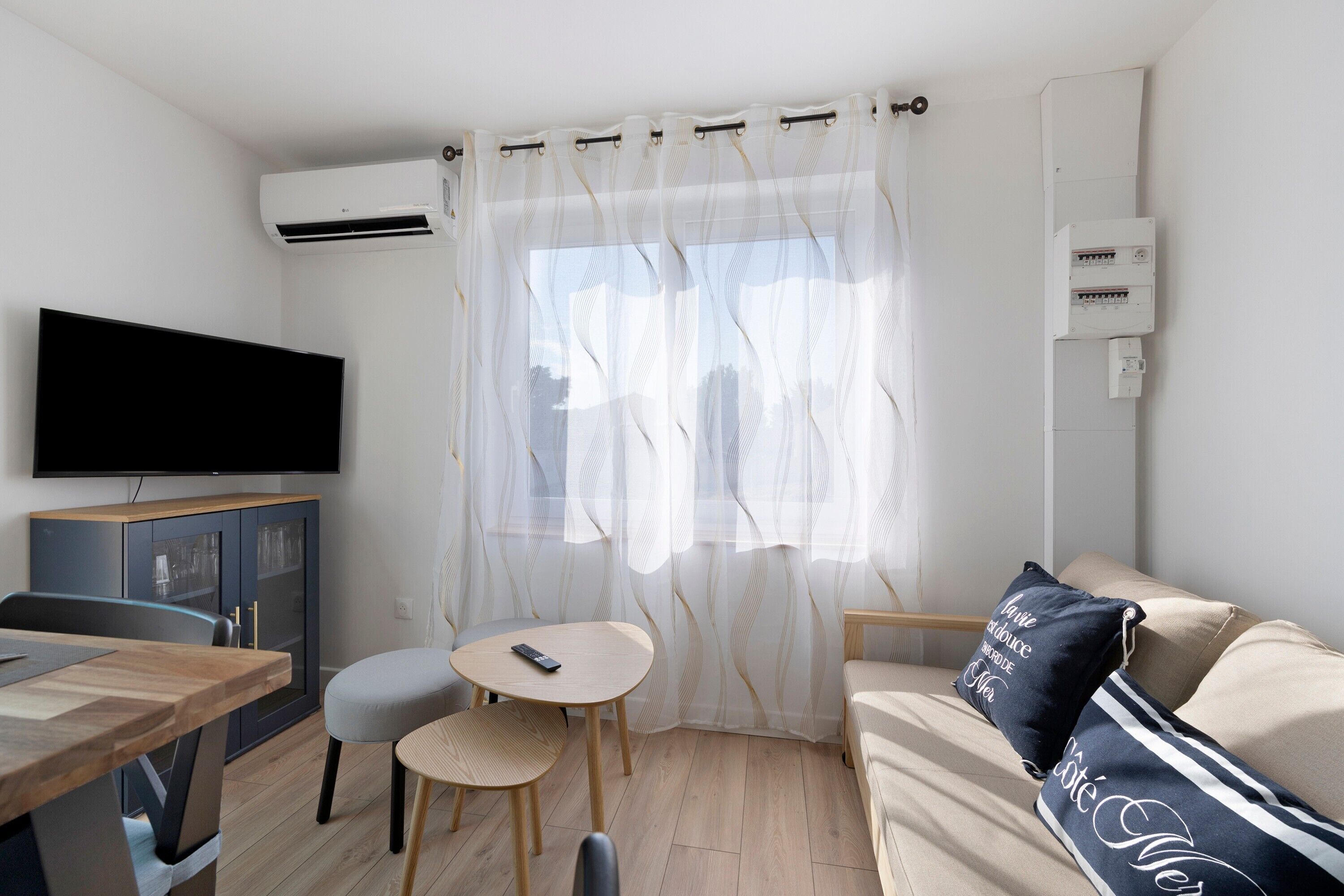 Una habitación con televisión de pantalla plana, una mesa de comedor con sillas, un sofá con un cojín y una ventana con cortinas traslúcidas.
