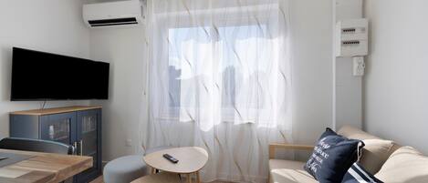 Una habitación con televisión de pantalla plana, una mesa de comedor con sillas, un sofá con un cojín y una ventana con cortinas traslúcidas.