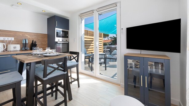 A modern kitchen with a dining area, a TV, and a sliding glass door leading to an outdoor seating area. - Holiday Home 'L'oeillet Bleu' with Private Terrace, Wi-Fi and Air Conditioning (Saint-Jean-de-Monts)