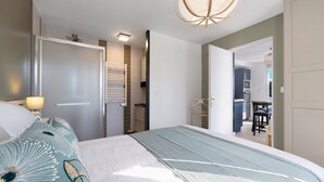 A hotel room with a bed, a bedside table with a lamp, a wardrobe, a bathroom with a towel rack, and a view into the kitchen area. - Holiday Home 'L'oeillet Bleu' with Private Terrace, Wi-Fi and Air Conditioning (Saint-Jean-de-Monts)