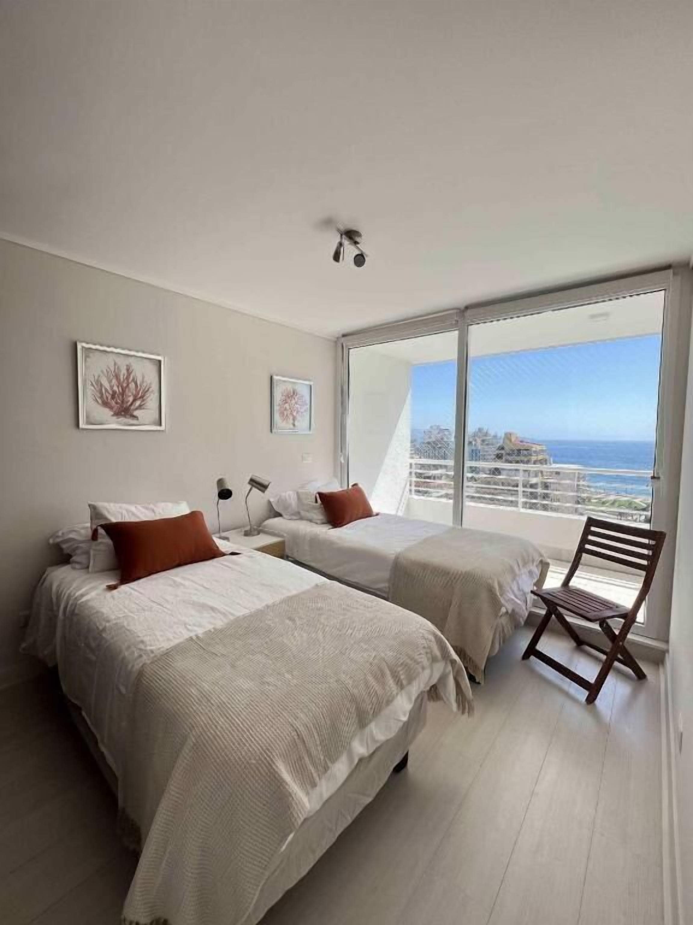 A bedroom with two beds, a chair, and a large window with a view of the sea.
