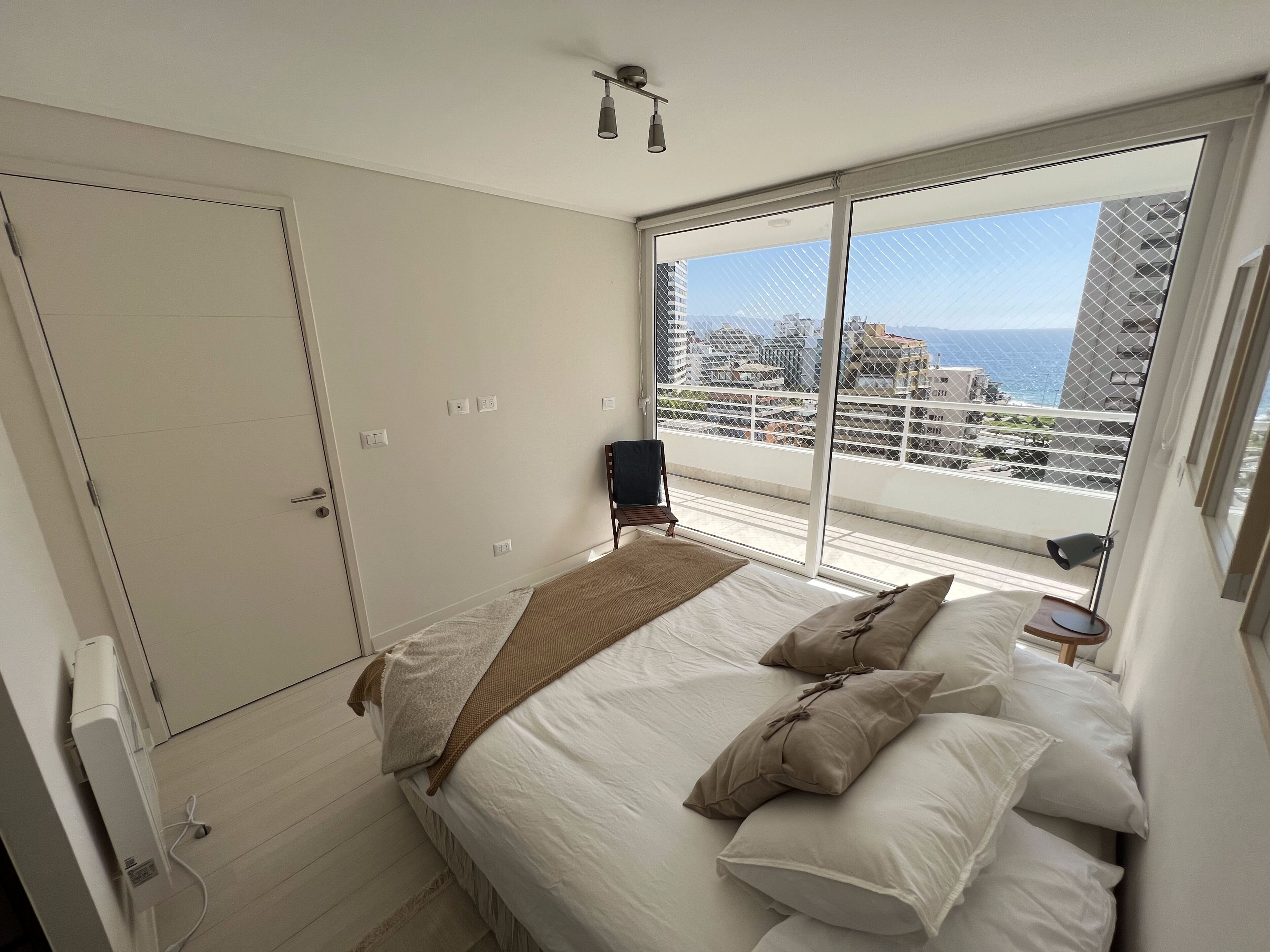 A bedroom with a bed, a chair, a window with a view of buildings and the sea, and a ceiling light.