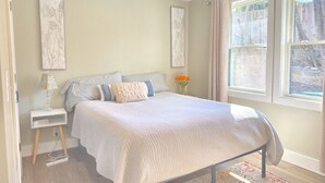 Ein Schlafzimmer mit einem Bett, einem Nachttisch, einer Lampe, einer Vase mit Blumen und einem Fenster mit Vorhängen.