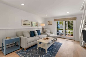 A living room with a sofa, a coffee table, a side table, and a lamp. There are framed pictures on the wall and a view of the outside through a glass door. - Hydrangea Lane Beach Cottage (Mashpee)