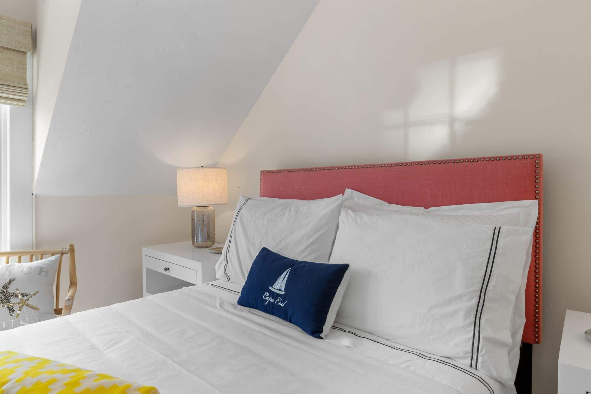 A bedroom with a white bed, a red headboard, a nightstand with a lamp, and a window with blinds.