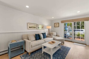 A modern living room with a sofa, coffee table, side table, and a view through large windows. - Hydrangea Lane Beach Cottage (Mashpee)
