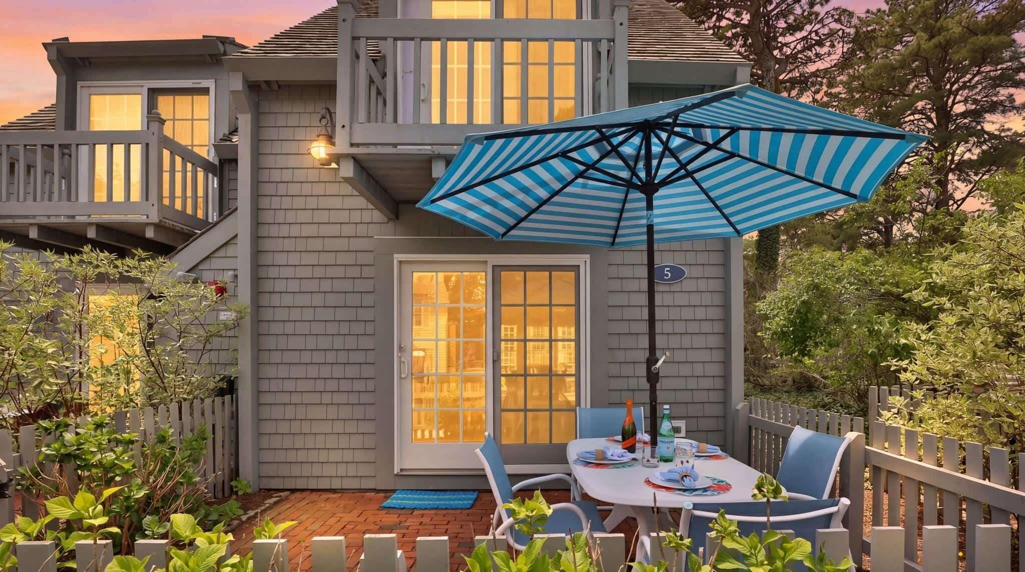 A patio with a table and chairs, a blue-striped umbrella, and a house with a grey exterior and a white fence.