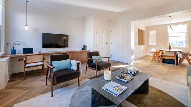 Ein modernes Wohnzimmer mit Flachbildfernseher, Holzmöbeln, einer Essecke mit Tisch und Stühlen sowie einem großen Fenster.