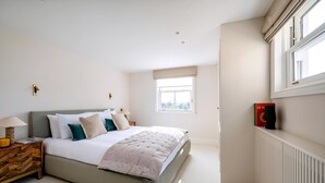 Ein Schlafzimmer mit Bett, Nachttisch, Lampe, Fenster, Heizkörper und einem an der Wand montierten Lautsprecher.