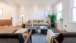 Ein modernes Wohnzimmer mit einer Couch, einem Couchtisch und Sesseln. Ein Heizkörper, eine Lampe und eine Pflanze sind ebenfalls vorhanden.