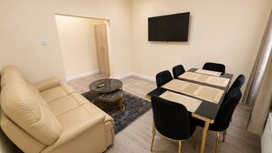Una sala de estar con un sofá color beige, una mesa redonda de centro, una mesa de comedor con sillas y un televisor de pantalla plana montado en la pared.