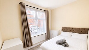 Un dormitorio con cama, radiador, ventana con cortinas y vistas a edificios.