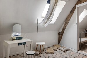 A bedroom with a vanity, a mirror, a small stool, and a bed with a metal frame. There are two windows providing natural light.