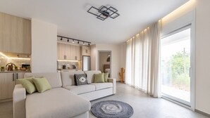 A modern living room with a sofa, a kitchen area, and a large window with curtains. - Enclave Luxury Apartments Zante Experience equiped with Jacuzzi,BBQ,GYM (Zakinthos)