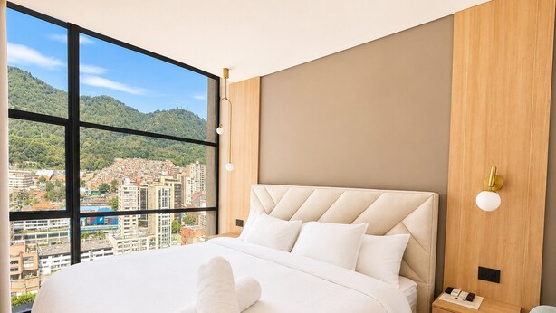 A modern hotel room with a large bed, a nightstand, a wall-mounted lamp, and a view of a cityscape through large windows. - King Bed + View Monserrate - 19 floor (Bogotá)