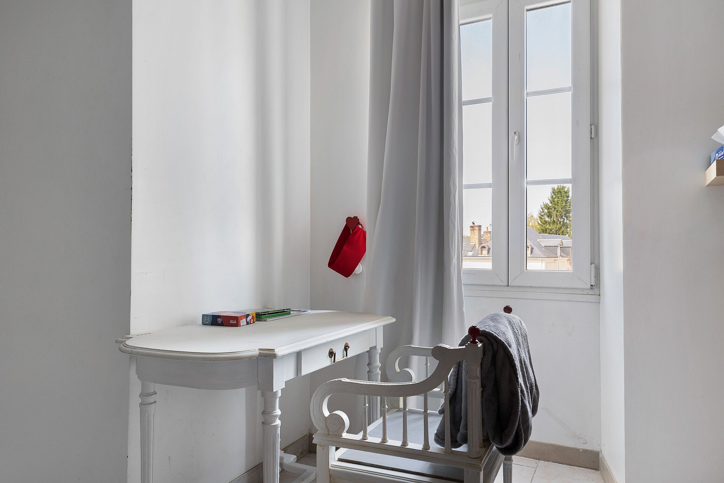 Ein Zimmer mit weißem Sessel, einem kleinen Tisch und einem Fenster mit Vorhängen.
