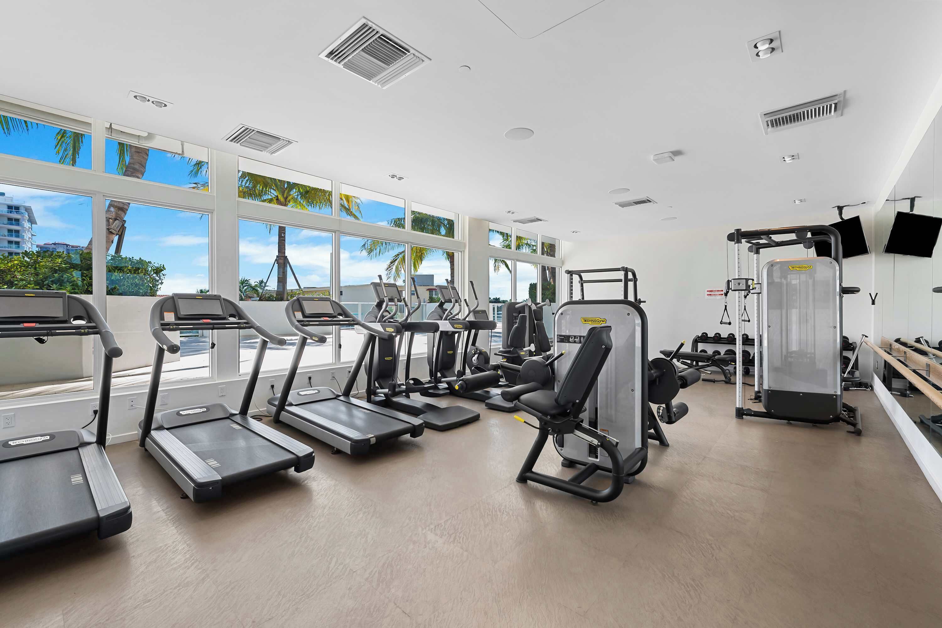 Une salle de sport équipée de tapis de course, d’appareils de musculation et dotée d’une grande fenêtre offrant une vue sur les palmiers et les bâtiments.
