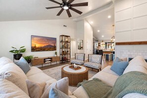 A spacious living room with a ceiling fan, a large painting, a wooden bookshelf, and a kitchen in the background. - NEW | Sonoran Sol |Modern Pool Oasis Near Kierland (Scottsdale)