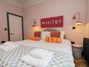 A hotel room with a bed, checkered bedspread, pillows, a wall sign, and wall-mounted lamps. - Apartment Pink (Whitby)