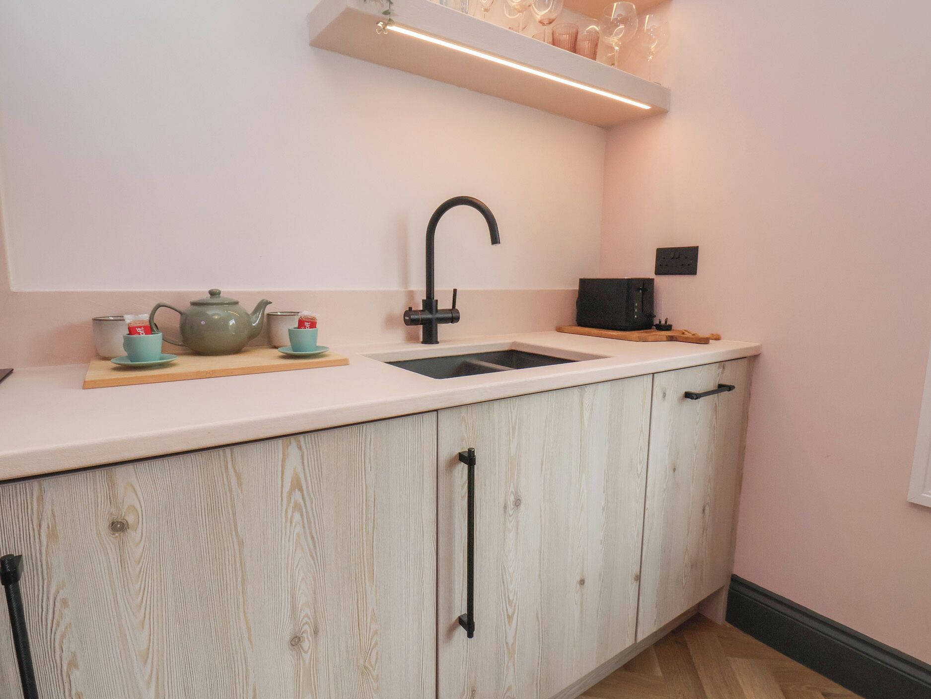 Una cocina con encimera blanca, grifo negro, armario de madera y repisa con cristalería.