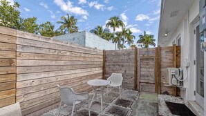 Un patio con valla de madera, dos sillas blancas, una mesa redonda pequeña y un cuadro de conexiones eléctricas exterior blanco.