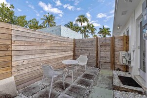 Un patio con valla de madera, dos sillas blancas, una mesa redonda pequeña y un cuadro de conexiones eléctricas exterior blanco.