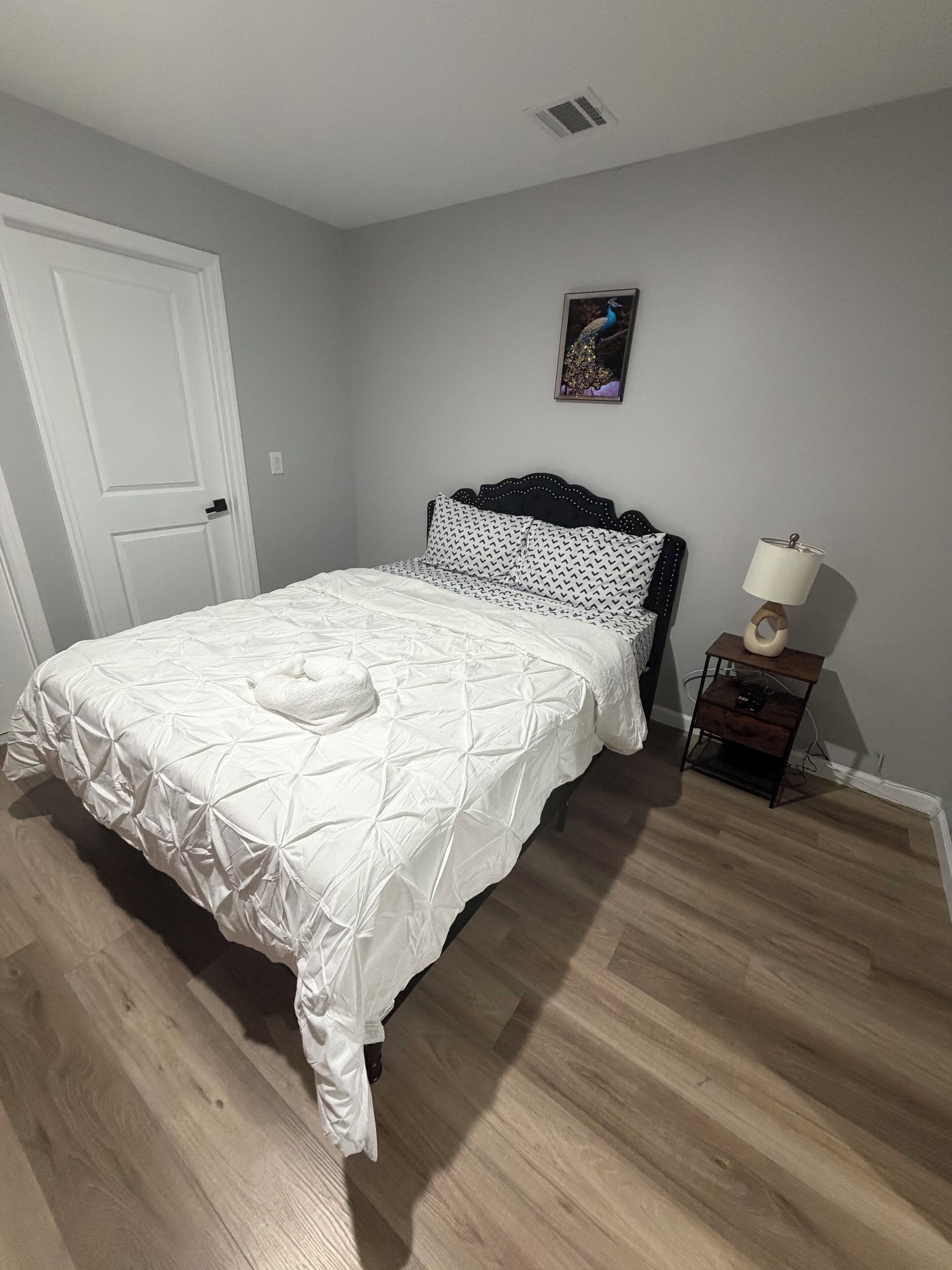 Ein Schlafzimmer mit Bett, Nachttisch, Lampe, einer Tür und einem an der Wand hängenden Bild.
