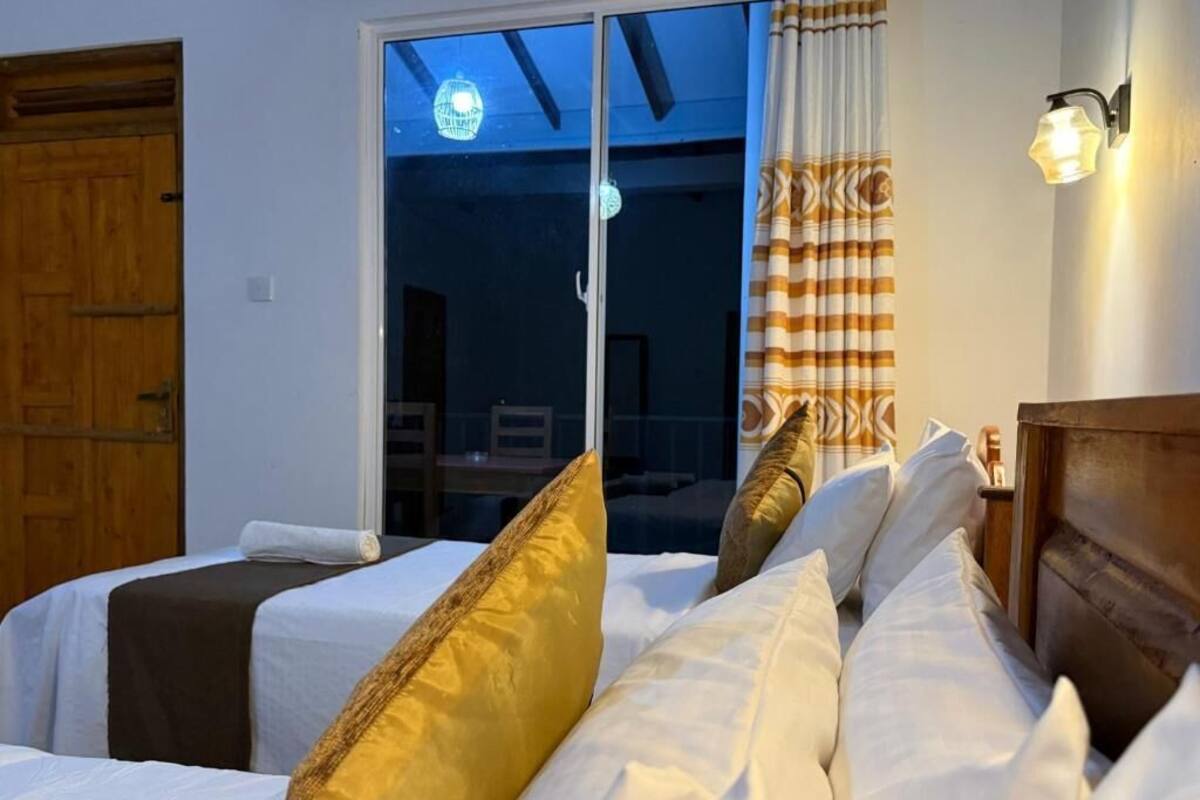 Habitación de hotel con cama, puerta de madera, ventana con cortinas y lámpara fijada a la pared.