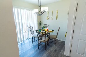 Un espace repas comprenant une table, des chaises, un lustre et une fenêtre avec des stores.