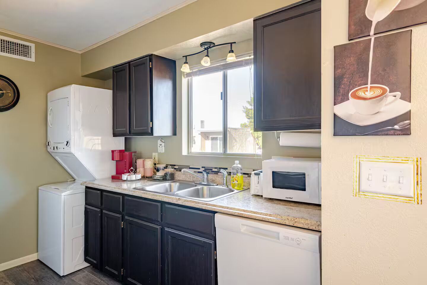 A kitchen with a dishwasher, sink, and a microwave. There is a window above the sink and a clock on the wall.