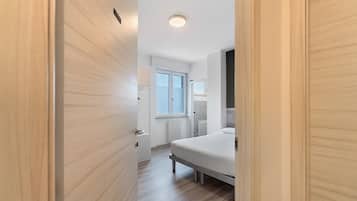 Ein schmaler Flur mit einer Holztür links, einem weißen Heizkörper und einem Bett mit weißer Matratze im Zimmer.