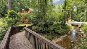 Eine Holzterrasse führt zu einer rustikalen Holzhütte, umgeben von dichtem Wald und einem kleinen Bach.