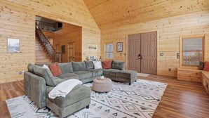 L’intérieur d’une cabane en rondins, avec un canapé d’angle, un pouf, un tapis à motifs, un plancher en bois et un escalier menant à un étage supérieur.