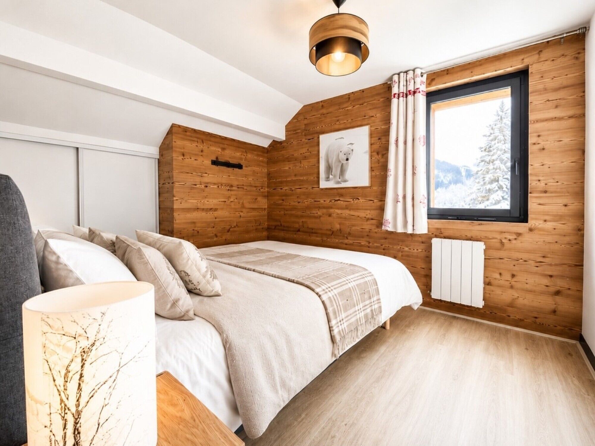 Ein Schlafzimmer mit Bett, einem Fenster mit Vorhängen, einer Holzwand und einer Deckenleuchte.