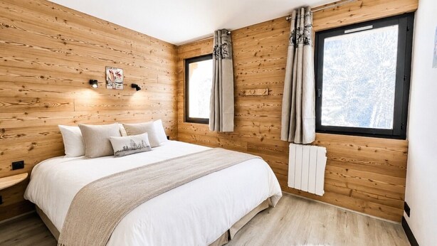 A bedroom with a wooden wall, a bed with white bedding, a fluffy rug, and a window with curtains. - Apartment - Le Batchu - Valloire - 12 guests (Valloire)