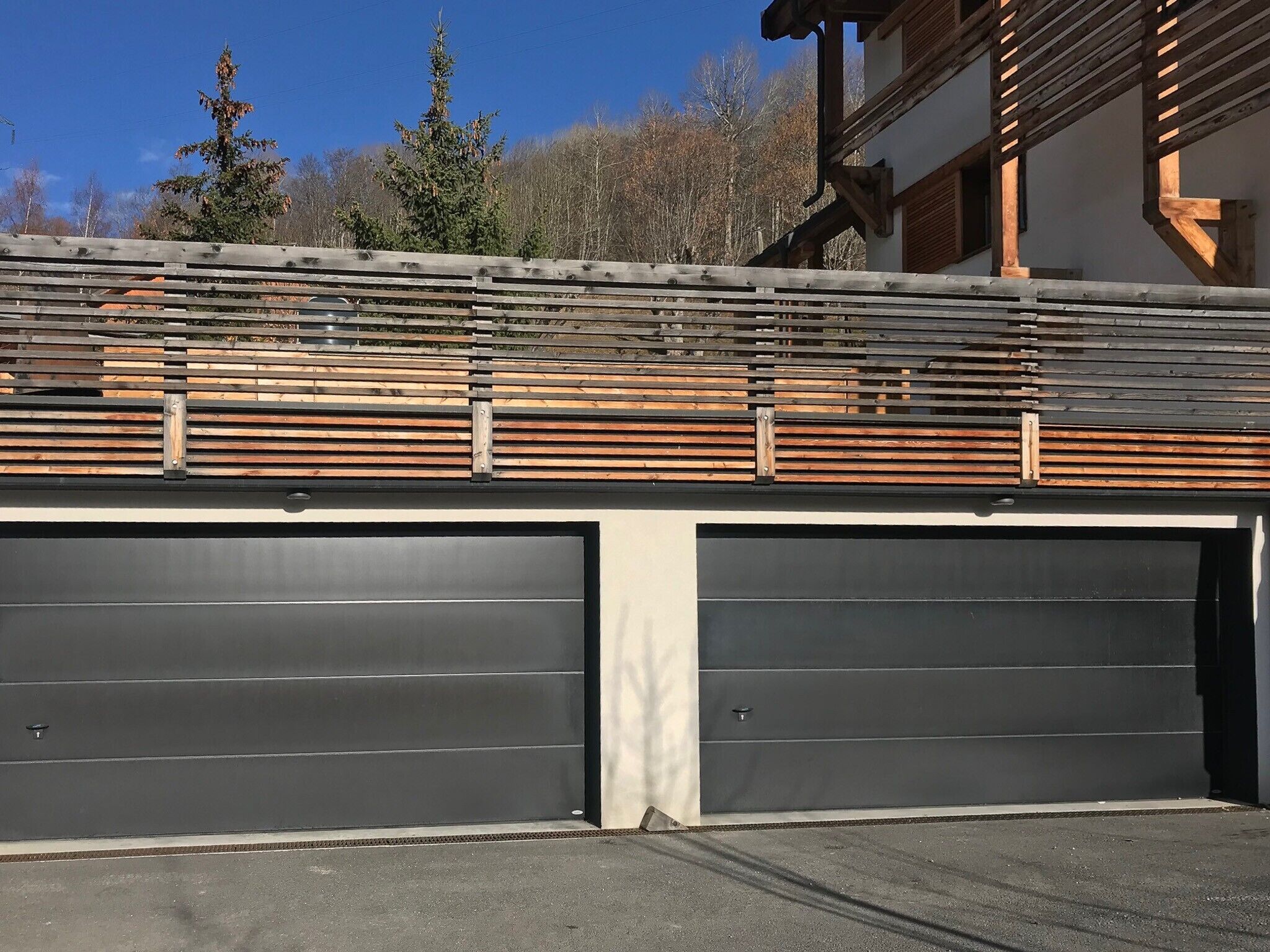 A garage with two large doors, a wooden fence, and a building with a sloped roof in the background.