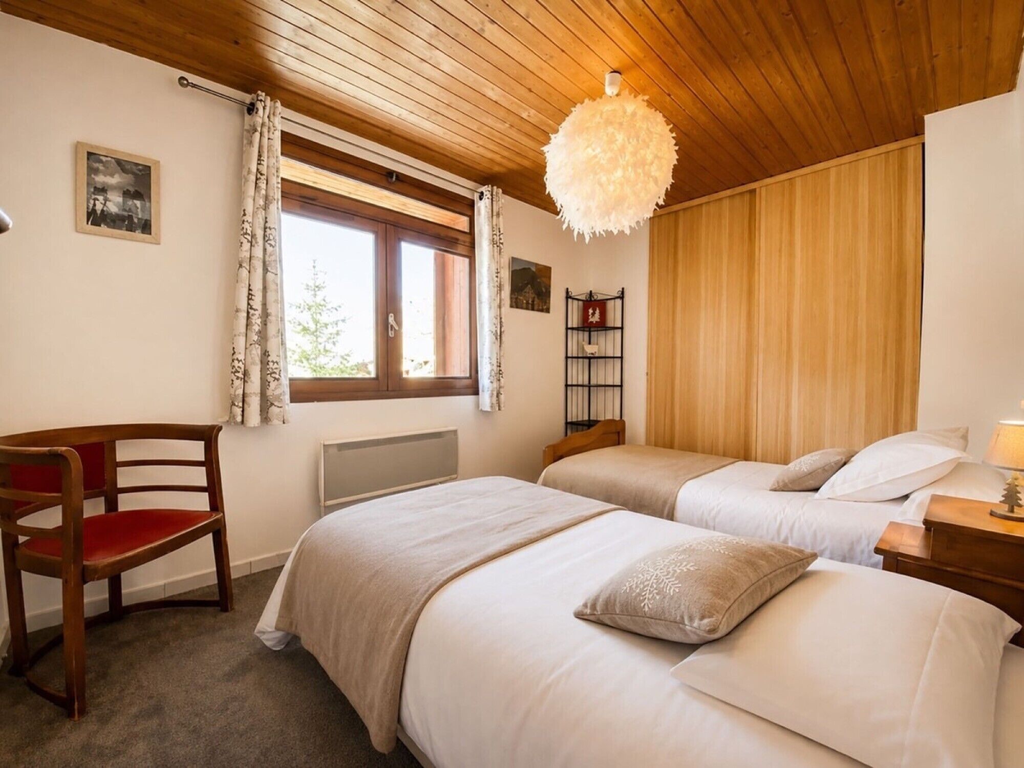 A room with two beds, a wooden chair, a radiator, a window with curtains, a hanging light fixture, and a shelf with a small tree decoration.