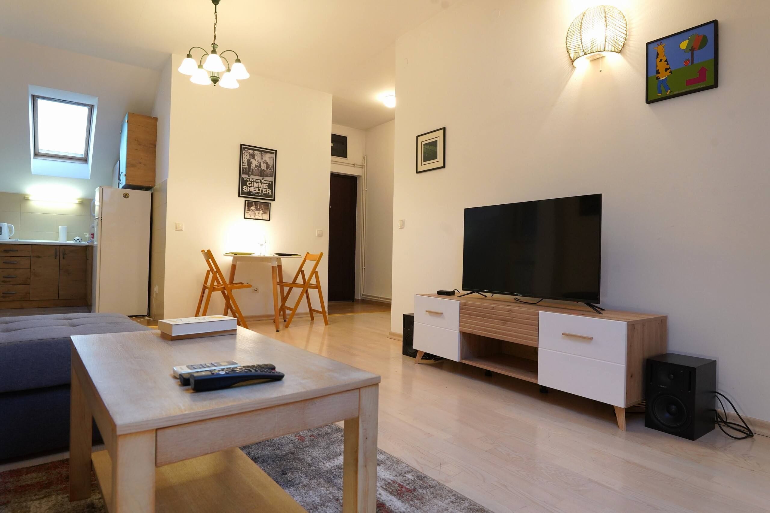 Uma sala de estar moderna com televisão, uma mesa de jantar e uma cozinha ao fundo.