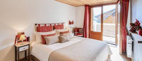 Un dormitorio con techo de madera, una cama grande con ropa de cama roja y blanca, una mesita de noche con una lámpara, una silla y una ventana con cortinas.