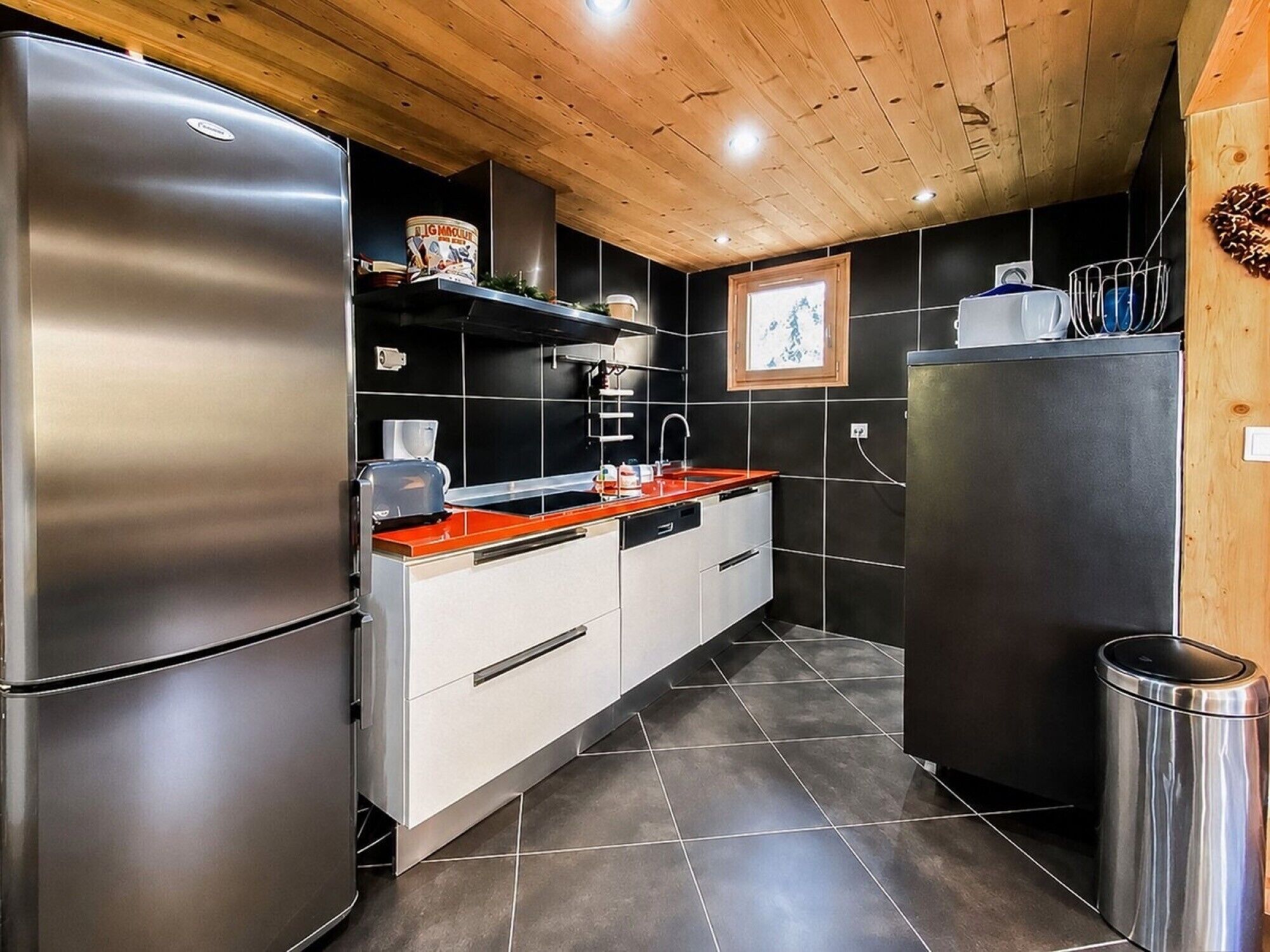 A modern kitchen with stainless steel appliances, black tiled walls, and wooden ceiling.