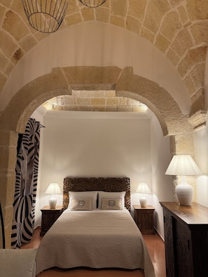 A bedroom with a stone archway, a bed with a quilted headboard, two bedside lamps, and a decorative lamp on a wooden table. - Angel Apartment 1 - Two Bedroom Apartment, Sleeps 4 (Monopoli)