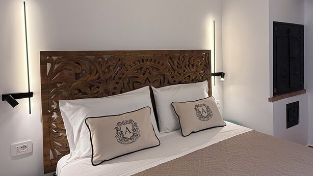 A bedroom with a wooden headboard, two pillows with monogrammed covers, a bedside lamp, and a wall-mounted TV. - Angel Apartment 1 - Two Bedroom Apartment, Sleeps 4 (Monopoli)