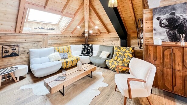 A cozy log cabin interior with a loft bedroom, wooden beams, a sofa set, a coffee table, and a bear painting. - Chalet Natura Lodge Valloire – Elegant Retreat for 10 Guests with Sauna and Mountain Views (Valloire)