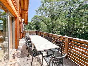 Un terrazza in legno con tavolo e sedie, circondata da un parapetto in legno, con vista su una zona boschiva.