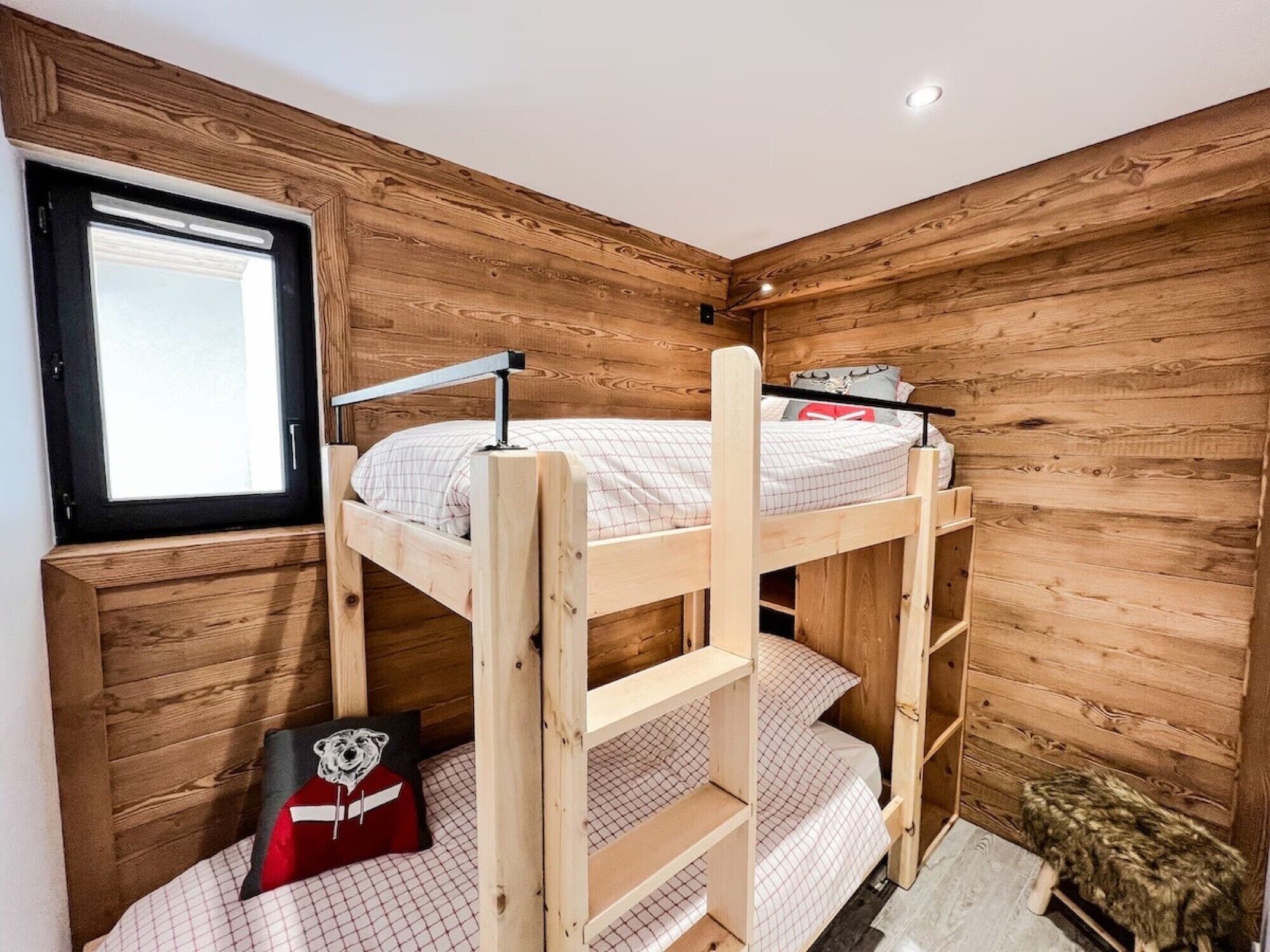 Habitación con literas, paredes de madera, una ventana y un pequeño banco.
