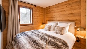 Un dormitorio de cabaña de madera con una cama, cortinas, una ventana y una mesita de noche con lámpara.